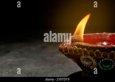 Ein rot-goldener Topf mit blauem und weißem Design steht auf einer dunklen Oberfläche. Im Topf brennt eine kleine Flamme. Diwali-Konzept Stockfoto