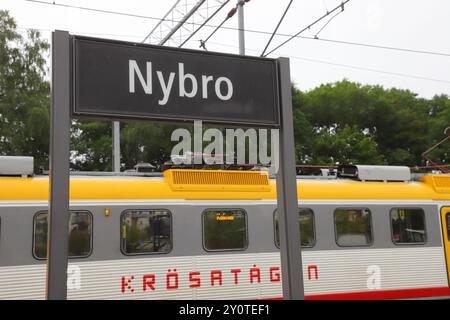 Nybro, Schweden - 8. August 2024: Nahaufnahme des Bahnhofsschilds von Nybro mit einem lokalen Personenzug im Hintergrund. Stockfoto