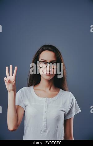 Eine junge Frau, die eine Brille trägt, hält drei Finger mit einem neutralen Gesichtsausdruck hoch. Sie steht vor einem blauen Hintergrund Stockfoto