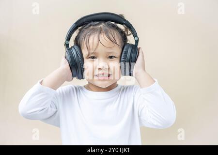 Ein glückliches asiatisches Mädchen im Vorschulalter, das ein weißes langärmeliges T-Shirt mit kabellosen Kopfhörern trägt und Musik hört oder Online-Unterricht auf einer Creme-Ba abspielt Stockfoto