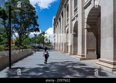 Hongkong, China - 03. Juli 2024 : Eine einsame Frau geht auf einem SteinGehweg vorbei an einem klassischen Gebäude mit einer Reihe von Säulen und Bögen. Stockfoto