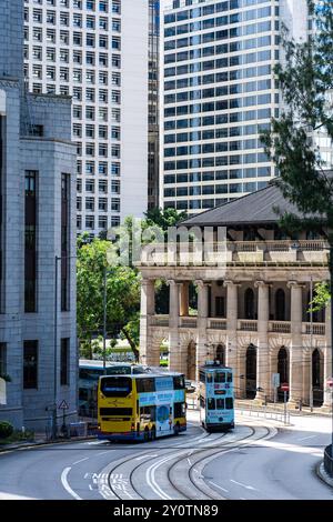 Hongkong, China - 03. Juli 2024 : Ein gelber Doppeldeckerbus und eine blaue Straßenbahn parken auf einer gekrümmten Straße vor einem klassischen Gebäude in Hong Kon Stockfoto