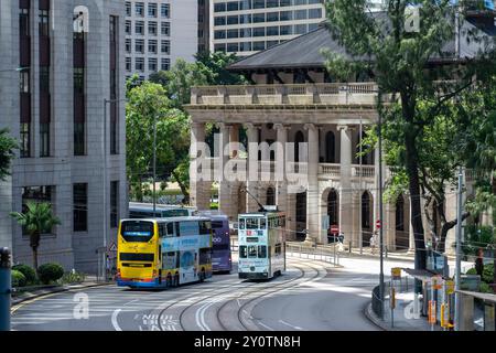 Hongkong, China - 03. Juli 2024 : Ein Doppeldeckerbus und eine Straßenbahn fahren entlang einer verwinkelten Straße in einer Stadt. Es gibt Hochhäuser auf der eithe Stockfoto