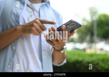 Zugeschnittene Aufnahme eines Mannes, der eine Textnachricht auf dem Mobiltelefon eingibt und im Freien steht Stockfoto