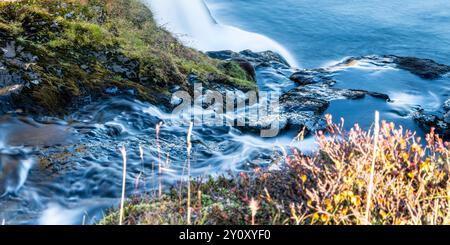 Ein Wasserstrom fließt über Felsen und Gras. Seideneffekt. Das Wasser ist ruhig und klar und die Felsen sind zerklüftet und rau. Das Gras ist hoch Stockfoto