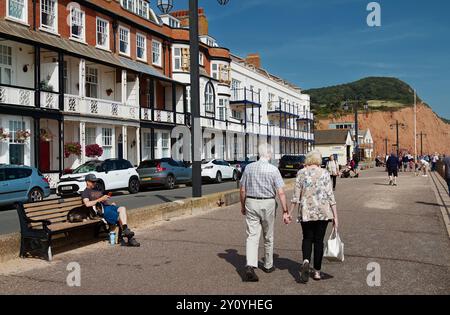 Blick entlang der Esplanade mit Öffentlichkeit, Menschen und Touristen, die zu den Red Cliffs of the Jurassic Coast, Sidmouth, Großbritannien, gehen und sitzen Stockfoto