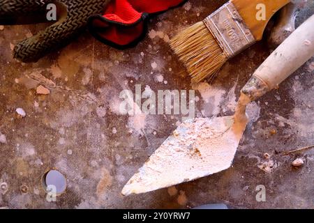 Ein Kontrast der Texturen zwischen den weichen Borsten eines Pinsels und der Rauheit des Mörtels auf einer Spitzkelle, die auf einem Maurerständer liegt. Stockfoto