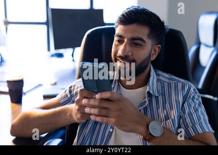 Mit Smartphone sitzt ein indischer Mann im Bürostuhl, lächelt und entspannt Stockfoto