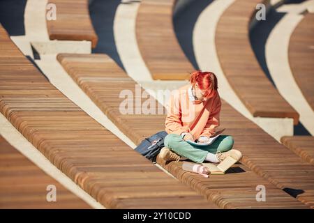 Studentin mit leuchtend roten Haaren im Notizbuch, die auf einer gekrümmten Amphitheater-Bank sitzt, mit offenem Buch und Schulrücken neben dem Sitz, lässige Outdoor-Lernszene, Kopierraum Stockfoto