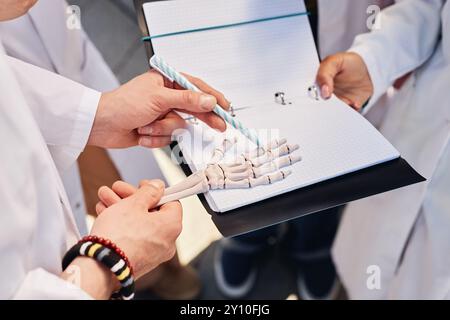 Nahaufnahme der Hände eines Medizinstudierenden, der das anatomische Modell eines menschlichen Handskeletts untersucht, das mit Stift auf die Knochen von Phalangen zeigt, während er im Kreis mit Klassenkameraden im Freien steht, Kopierraum Stockfoto