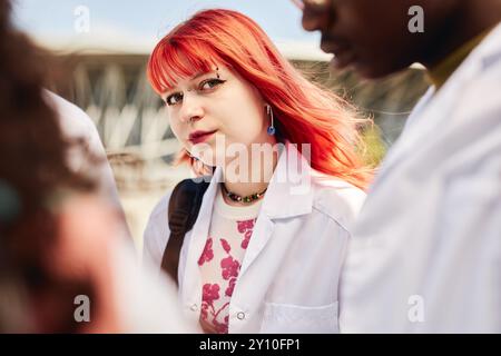 Porträt einer jungen Medizinstudentin mit leuchtend roten Haaren, die lächelnd und in die Kamera blickend, während sie mit Klassenkameraden draußen an sonnigen Tagen im Kopierraum steht Stockfoto