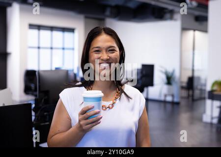 Lächelnde Frau, die im modernen Büro eine Kaffeetasse hält und die Pause genießt Stockfoto