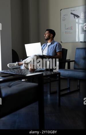 Er arbeitet am Laptop, ein indischer Mann sitzt in einem modernen Büro mit Whiteboard im Hintergrund, Kopierraum Stockfoto