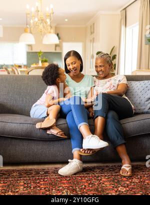 Auf der Couch sitzen drei Generationen von Frauen, die zu Hause lachen und knüpfen Stockfoto