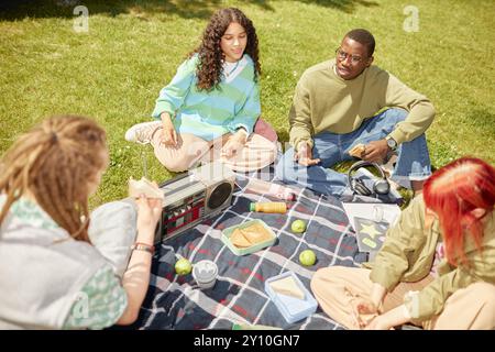 Verschiedene Gruppen von Studenten treffen sich zum Picknick auf dem grünen Rasen, unterhalten sich und genießen sich in Gesellschaft, während sie bei Sonnenschein im Freien Snacks essen Stockfoto