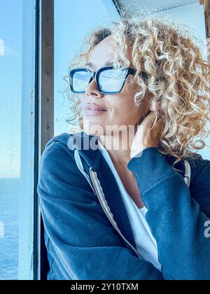 Schöne Frau mit Brille, die Freizeit mit Blick auf das Meer verbringt. Sie kümmert sich um sich, entspannt. Genießen Sie die Aussicht, schauen Sie durch das Fenster. Blonde und lockige Frauen mittleren Alters genießen die Aussicht. Ruhiger Morgen. Invia Commenti Stockfoto