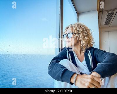 Schöne Frau mit Brille, die Freizeit mit Blick auf das Meer verbringt. Sie kümmert sich um sich, entspannt. Genießen Sie die Aussicht, schauen Sie durch das Fenster. Blonde und lockige Frauen mittleren Alters genießen die Aussicht. Ruhiger Morgen. Invia Commenti Stockfoto