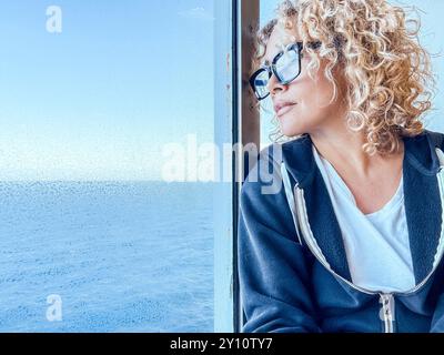 Schöne Frau mit Brille, die Freizeit mit Blick auf das Meer verbringt. Sie kümmert sich um sich, entspannt. Genießen Sie die Aussicht, schauen Sie durch das Fenster. Blonde und lockige Frauen mittleren Alters genießen die Aussicht. Ruhiger Morgen. Invia Commenti Stockfoto