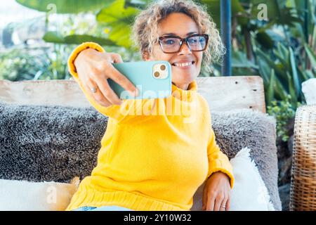 Eine Frau benutzt ihr Handy und entspannt sich allein im Garten auf einem Sofa. Porträt von fröhlicher weiblicher Schrift auf Handy. Frau macht Selfie. Reife junge Leute, die sich entspannen Stockfoto