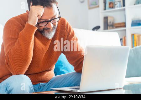 Trauriger und frustrierter Mann, der sich müde fühlt und besorgt über ein Problem, zu Hause mit Laptop auf dem Sofa zu sitzen, gestresster 50-jähriger Mann, der Probleme hat, schlechte Nachrichten online zu lesen, E-Mail-Benachrichtigung über Schulden oder negative Nachrichten Stockfoto