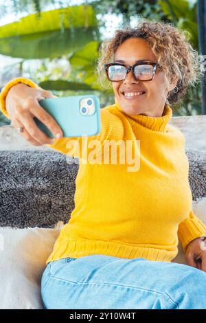 Eine Frau benutzt ihr Handy und entspannt sich allein im Garten auf einem Sofa. Porträt von fröhlicher weiblicher Schrift auf Handy. Frau macht Selfie. Reife junge Leute, die sich entspannen Stockfoto