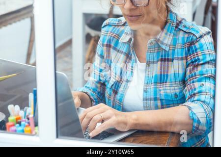 Frau in kariertem Hemd, die am Laptop arbeitet, während sie am Schreibtisch in der Nähe des Fensters im Labor oder zu Hause sitzt. Sicht durch das Fenster Stockfoto