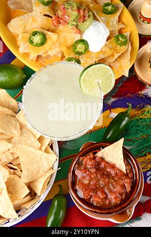 Blick von oben auf einen Margarita-Cocktail, umgeben von Nachos, Chips, Salsa auf einem hellen mexikanischen Tischtuch. Vertikales Format. Cinco de Mayo-Thema. Stockfoto