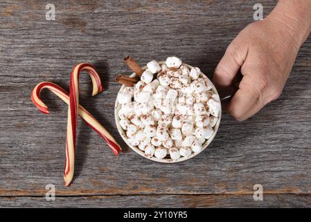 Blick von oben auf Santa Claus Hand mit einer großen Tasse heißen Kakao mit Marshmallows und Zimt. Zwei gekreuzte Süßigkeiten stehen neben dem Becher. Stockfoto