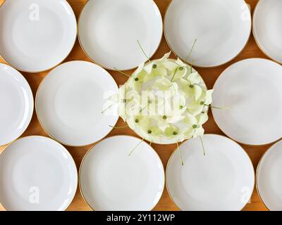 Ein weißer Teller mit goldenem Rand ist festlich mit den Blüten der Hartholzpflanze verziert, die anderen Teller sind leer, Vorbereitung und Dekoration für eine Feier im Sommer Stockfoto