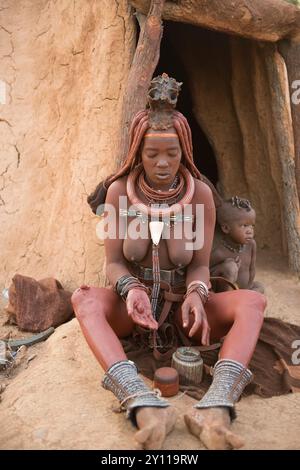 Junge Himba-Frau mit Kind auf der Morgentoilette vor der Hütte, wo die Frau ihre Haut mit einer Creme aus Butterfett, Steinmehl und ockerfarbiger Farbe reibt, Kaokoveld, Namibia Stockfoto