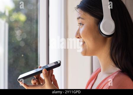 Sie trägt Kopfhörer, asiatische Frau verwendet Smartphone für Sprachbefehle in der Nähe des Fensters Stockfoto