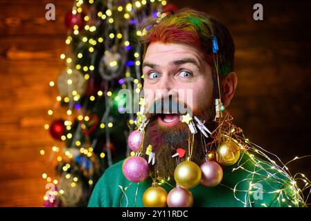 Weihnachtsfriseur. Lächelnder bärtiger Mann mit verziertem Bart. Weihnachtsdekoration. Weihnachtskugeln für Bartdekorationen. Ein schöner Mann, der sich auf die nächste vorbereitet Stockfoto