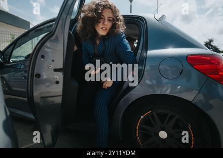 Geschäftsfrau verlässt das Auto in urbaner Umgebung mit lockigen Haaren in blauem Nadelstreifenanzug Stockfoto