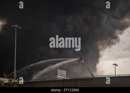 Ein Feuerwehrmann sprüht Wasser von einer hydraulischen Leiter, um einen großen Industriebrand zu löschen. Dicker schwarzer Rauch weht in den Himmel Stockfoto