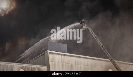 Ein Feuerwehrmann sprüht Wasser von einer hydraulischen Leiter, um einen großen Industriebrand zu löschen. Dicker schwarzer Rauch weht in den Himmel Stockfoto