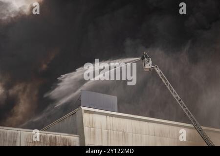 Ein Feuerwehrmann sprüht Wasser von einer hydraulischen Leiter, um einen großen Industriebrand zu löschen. Dicker schwarzer Rauch weht in den Himmel Stockfoto