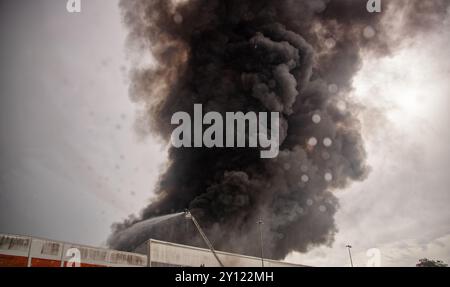 Ein Feuerwehrmann sprüht Wasser von einer hydraulischen Leiter, um einen großen Industriebrand zu löschen. Dicker schwarzer Rauch weht in den Himmel Stockfoto