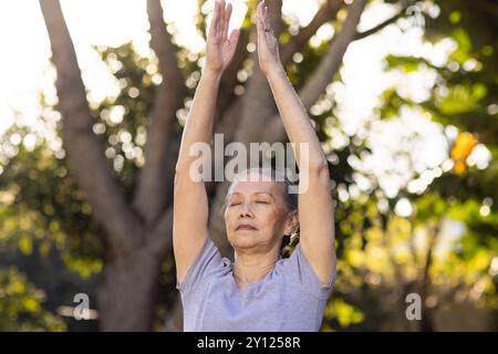 Yoga im Freien üben, ältere asiatische Frau die Arme hochhebt und die Augen schließt, die Natur genießt Stockfoto