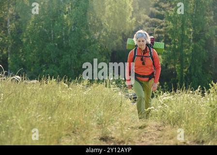 Reife Wanderer in Aktivkleidung und Stirnband, die einen Rucksack tragen, während sie den Waldweg entlang, umgeben von grünem Gras, befahren Stockfoto