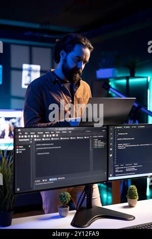 IT-Profis arbeiten von RGB-beleuchtetem Zuhause aus mit Laptops, testen und implementieren von Programmen und Systemen. Software-Techniker überprüft den Code, der auf Computermonitoren angezeigt wird, und steht in der Wohnung Stockfoto