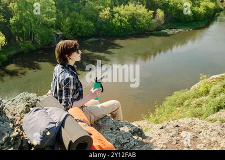 Junge Brünette weibliche Camper mit einer Flasche Wasser, die auf Klippen am Fluss sitzt und die Ruhe und die wunderbare Landschaft genießt Stockfoto