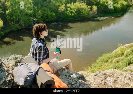 Junge Brünette weibliche Camper mit einer Flasche Wasser, die auf Klippen am Fluss sitzt und die Ruhe und die wunderbare Landschaft genießt Stockfoto