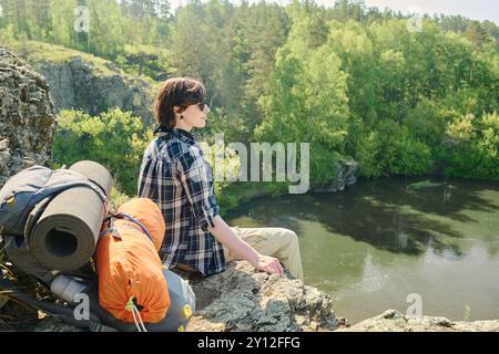 Junge, erholsame Wanderer in kariertem Hemd und Sonnenbrille, die auf dem Gipfel des Berges sitzen und während der Fahrt den Fluss bewundern Stockfoto