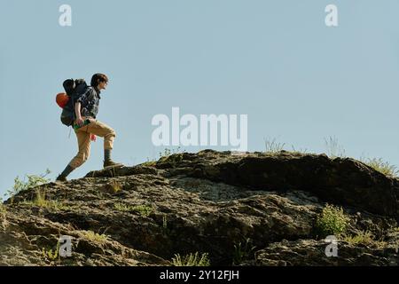 Lange Aufnahme einer jungen aktiven Frau in lässiger Kleidung, die einen Rucksack trägt, während sie den Gipfel des Berges gegen den blauen Himmel hinunterzieht Stockfoto
