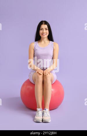 Sportliche junge Frau, die auf Fitball vor Fliederhintergrund sitzt Stockfoto