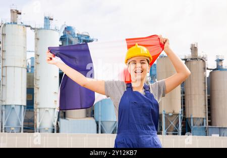 Positive junge Mitarbeiterin im Helm, die an einem sonnigen Sommertag vor großen Panzern in der Raffineriefabrik vor der französischen Nationalflagge schwenkt Stockfoto