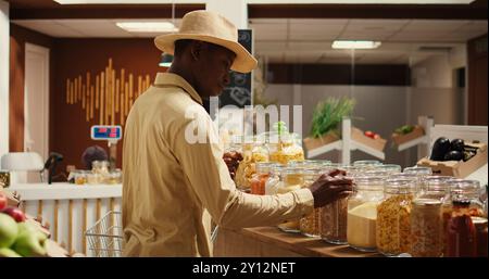 Der Kunde untersucht Getreide und Getreide im Zero Waste Öko Store und sucht nach natürlichen Bio-Produkten und Speiseküchen. afroamerikaner kauft hausgemachte Gewürze und Nudeln in Gläsern. Kamera 2. Stockfoto