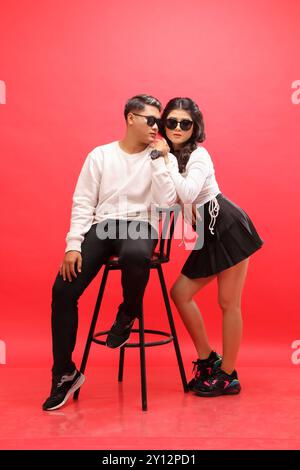 Langes junges Paar eleganter asiatischer Mann und Frau mit weißen Pullover, Sonnenbrille Mann, der seitlich auf einem Stuhl sitzt, während die Frau sich beugt Stockfoto