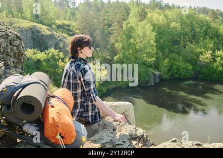 Junge, erholsame Wanderer in kariertem Hemd und Sonnenbrille, die auf dem Gipfel des Berges sitzen und während der Fahrt den Fluss bewundern Stockfoto