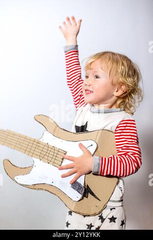 Ein fröhliches kleines Kind mit lockigen Haaren hebt die Hand, während es eine Pappgitarre hält. Mit gestreiftem Pyjama versprühen sie Spannung und Kreativität Stockfoto